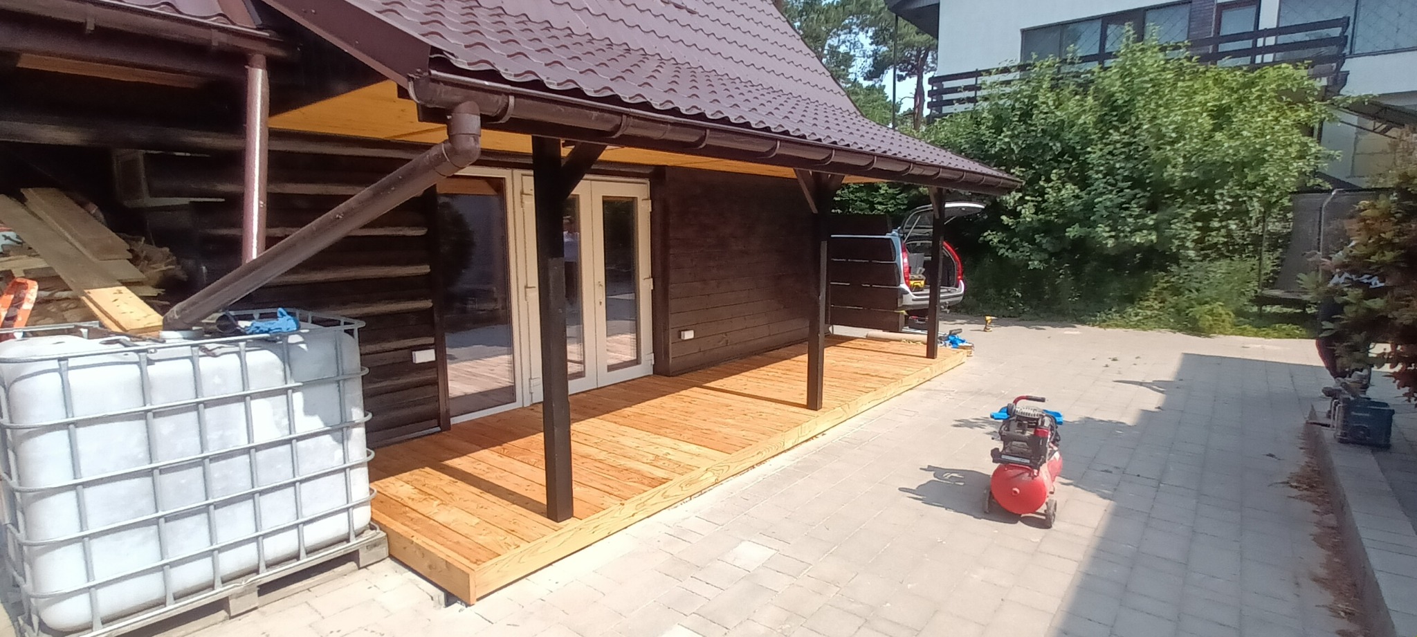 Nowo wybudowany drewniany taras przy domku letniskowym z brązowym dachem, widoczny kompresor, zbiornik na wodę i otwarty bagażnik samochodu z narzędziami.