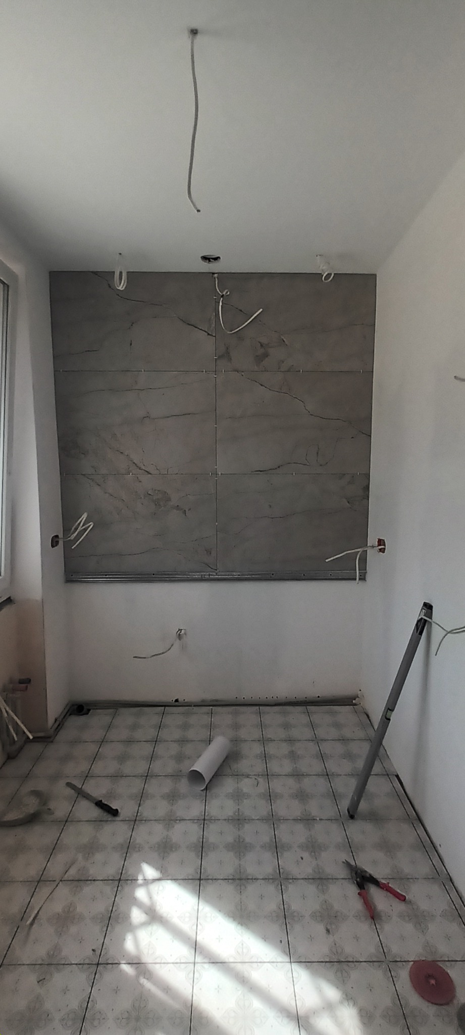 Wnętrze w trakcie remontu: położone płytki imitujące marmur na ścianie, wystające przewody elektryczne, narzędzia na podłodze z kwadratowych płytek w kwiatowy wzór, widoczne profile i rura z tworzywa.