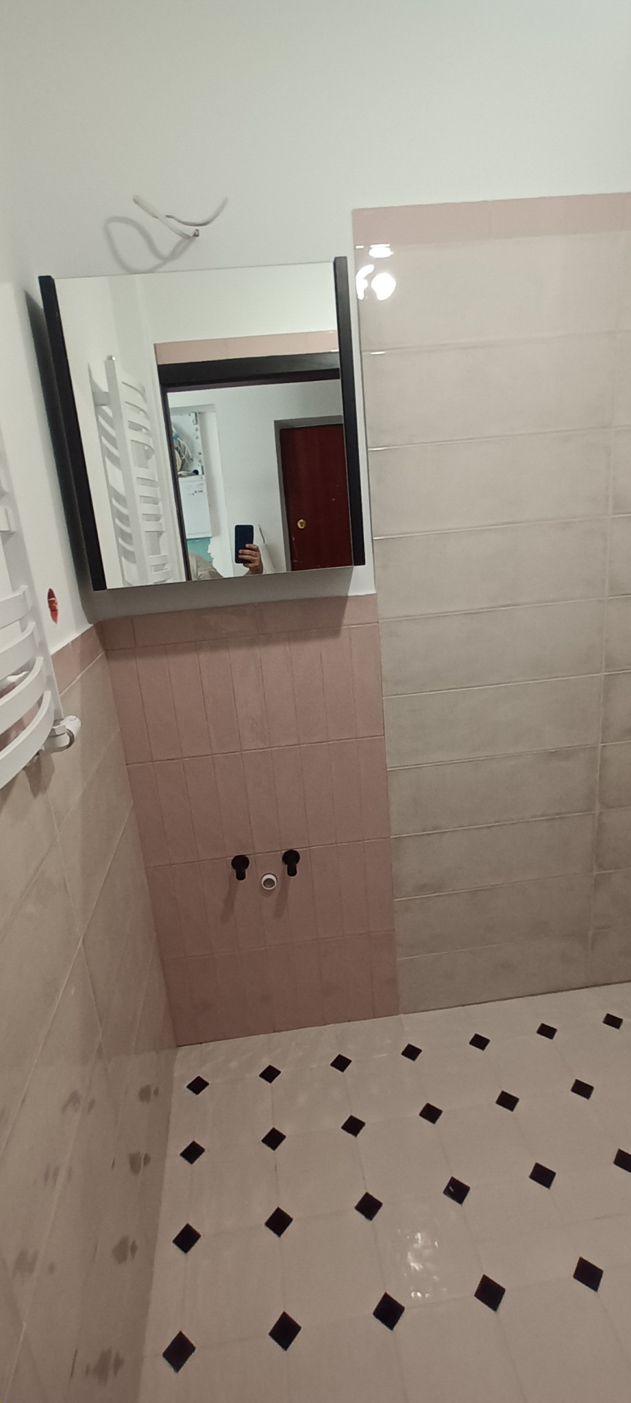 Łazienka w trakcie remontu: widoczne płytki ścienne w odcieniach różu i szarości, podłoga w biało-czarne romby, niezamaskowane rury przyłączeniowe do umywalki oraz lustro z czarną ramą, w którym...