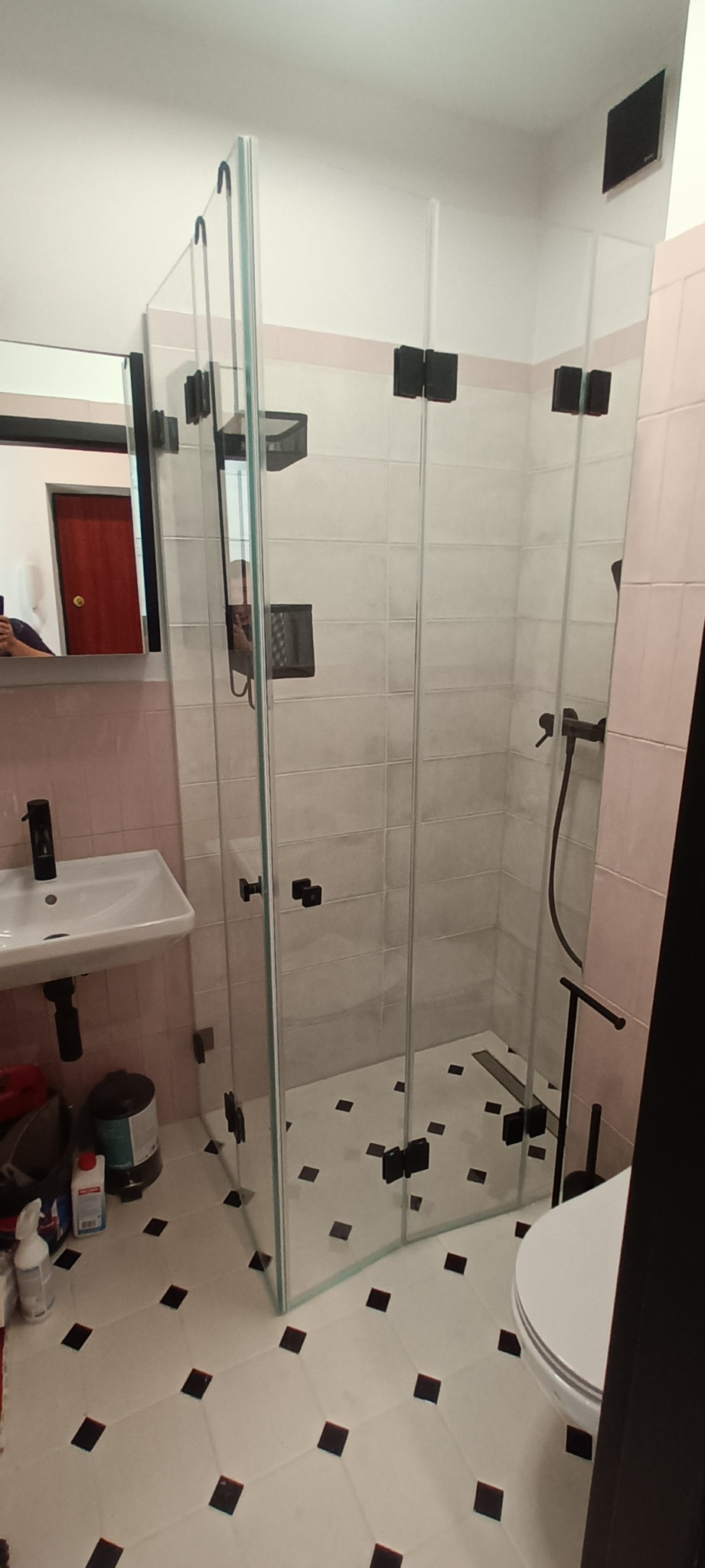 Nowoczesna kabina prysznicowa z czarnymi okuciami, białe płytki z czarnymi wstawkami na podłodze, umywalka z czarną baterią, różowe płytki na ścianach, widoczne akcesoria łazienkowe.