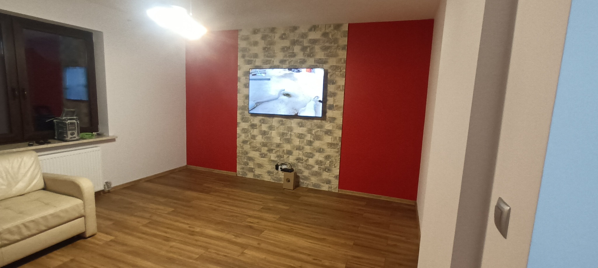 Salon po remoncie z panelami podłogowymi, ścianą w odcieniach czerwieni i szarości, oraz telewizorem na ścianie. Widoczny fragment sofy w lewym dolnym rogu.