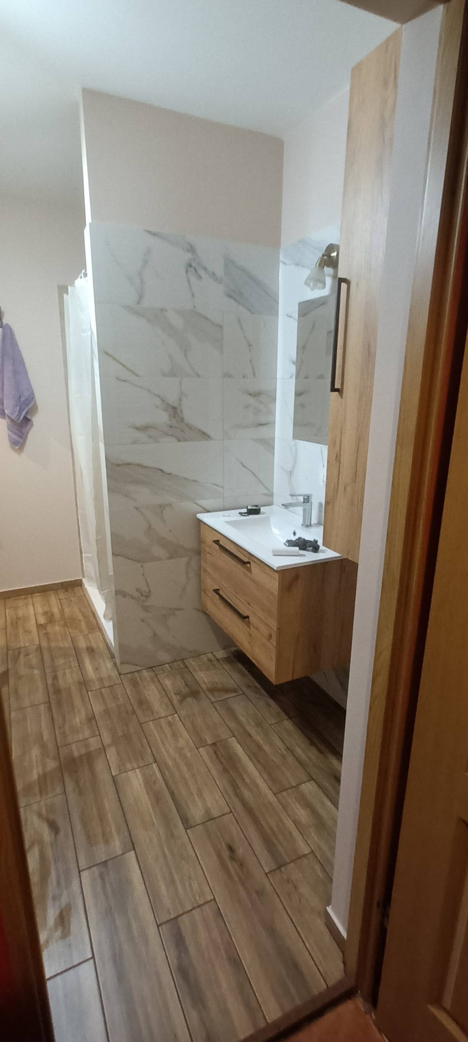 Wykończona łazienka w Łochowie z płytkami imitującymi marmur na ścianach, drewnopodobną podłogą i szafką pod umywalką z drewnianymi frontami.