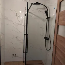 Kabina prysznicowa typu walk-in z czarną armaturą i marmurowymi płytkami na ścianach, drewniane drzwi po prawej stronie.