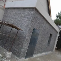 Wykończona elewacja budynku pokryta szarą, nieregularną okładziną kamienną, widoczne drzwi i okno, obok rusztowanie, teren wokół jeszcze nieuporządkowany.