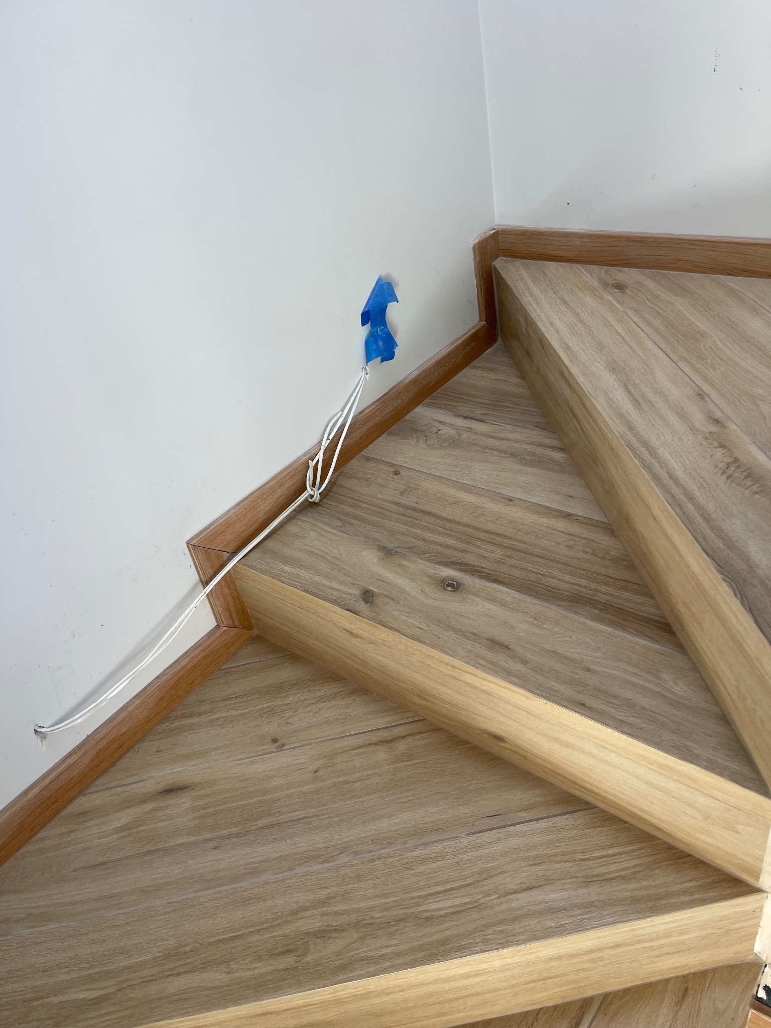 Schody wykończone panelami imitującymi drewno, z widocznym narożnikiem i wystającymi kablami zabezpieczonymi niebieską taśmą malarską przy ścianie.