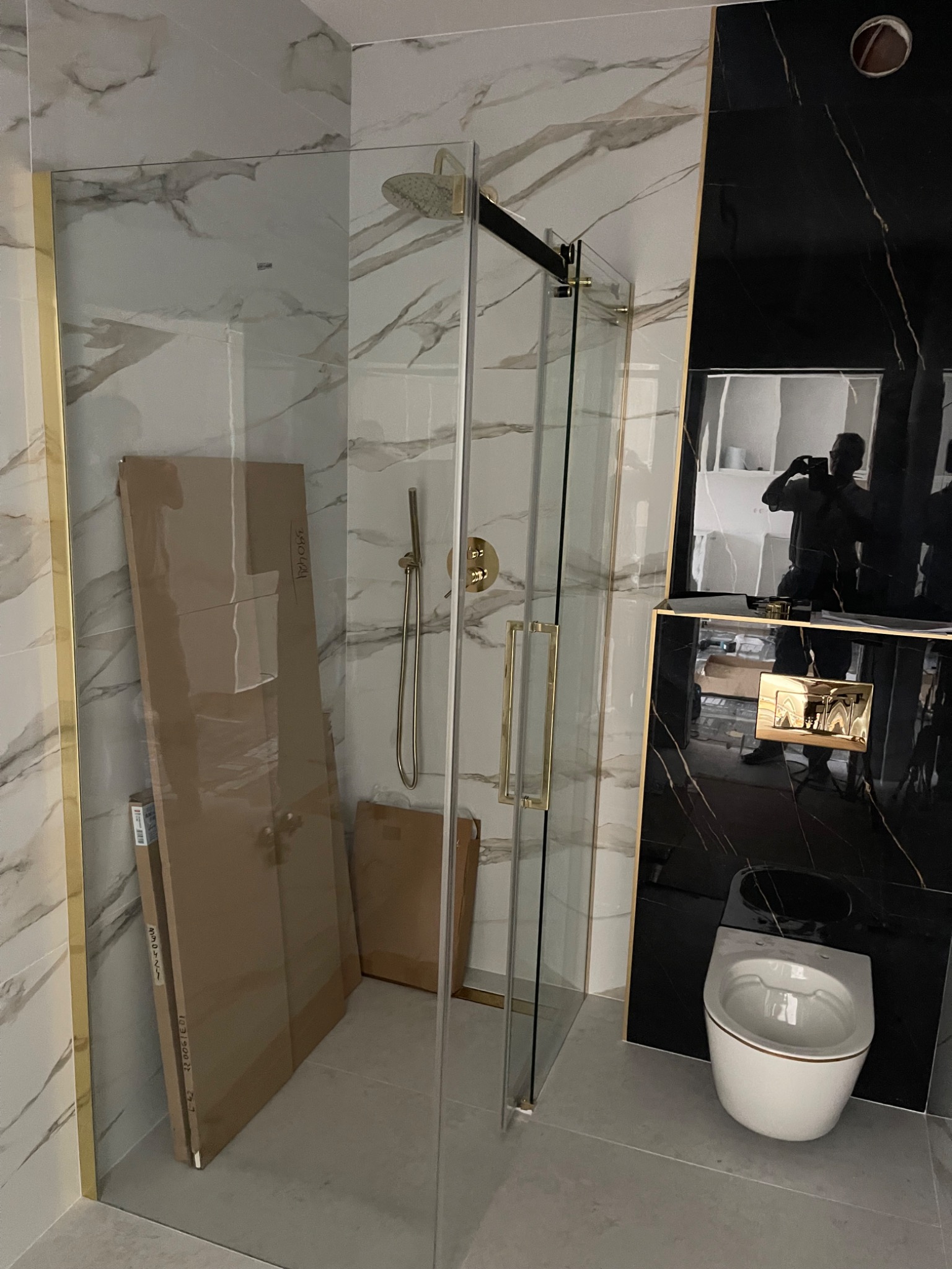 Nowoczesna łazienka w trakcie wykończenia: kabina prysznicowa ze złotymi elementami, marmurowe i czarne płytki, wisząca toaleta oraz kartony z panelami opartymi o ścianę.