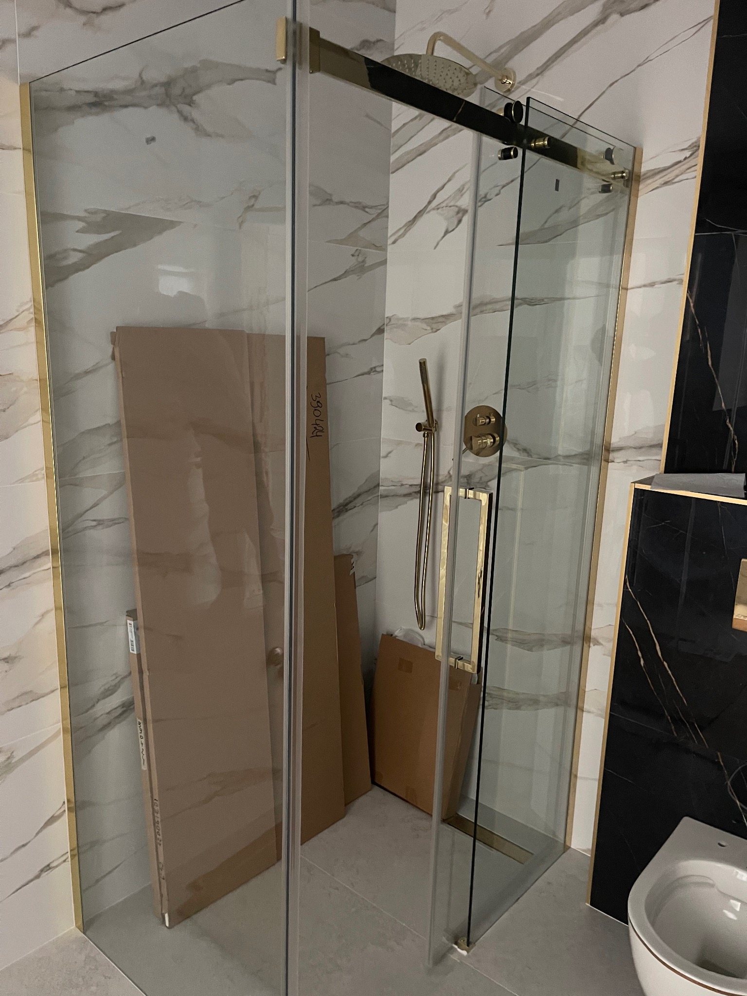 Szklana kabina prysznicowa z złotymi wykończeniami, zamontowana w łazience z marmurowymi płytkami, widoczne kartony z materiałami budowlanymi w tle, fragment ceramiki sanitarnej po prawej.
