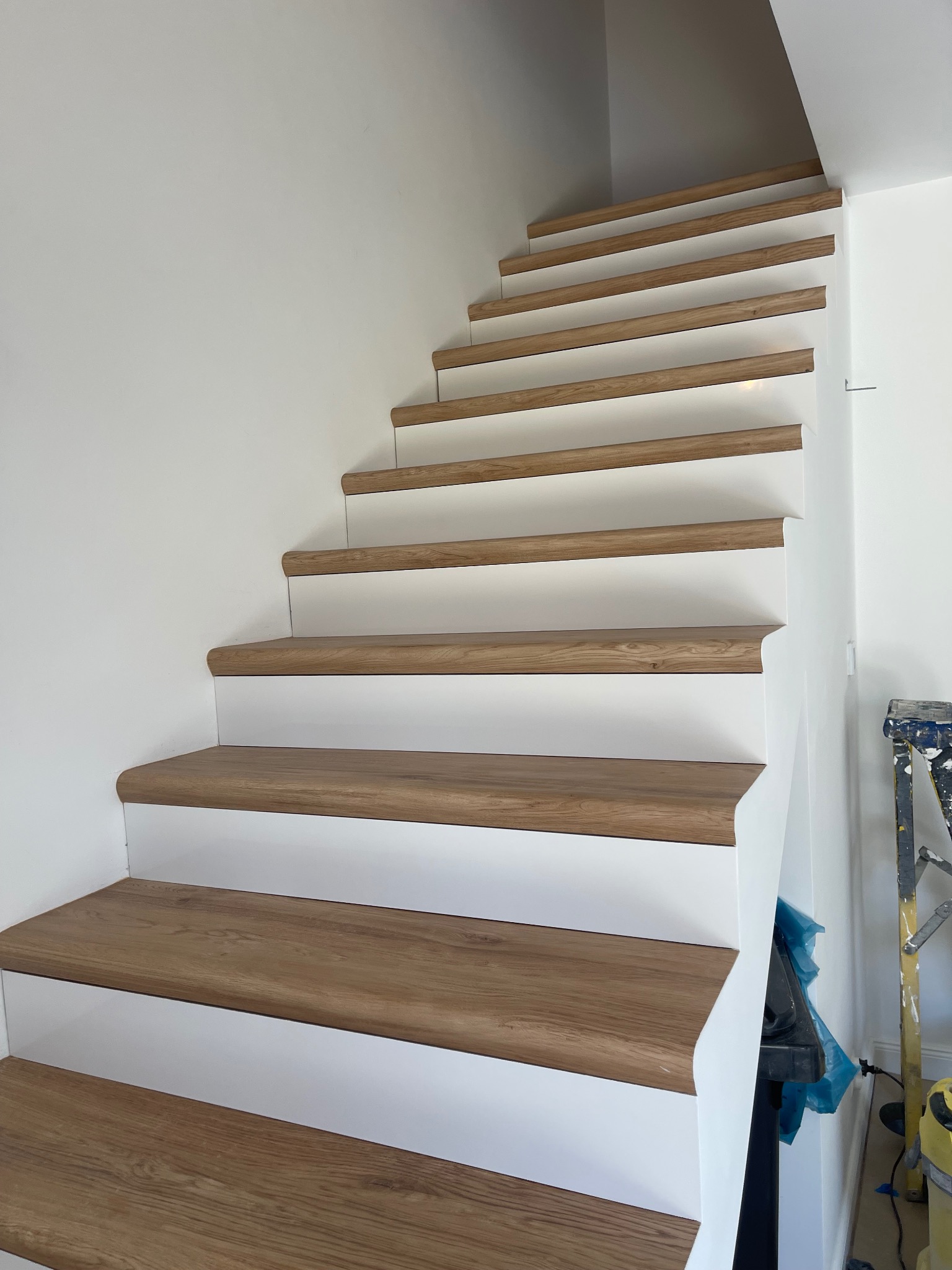 Schody z drewnianymi stopniami i białymi podstopnicami, widoczne z dołu, z fragmentem drabiny malarskiej w tle.