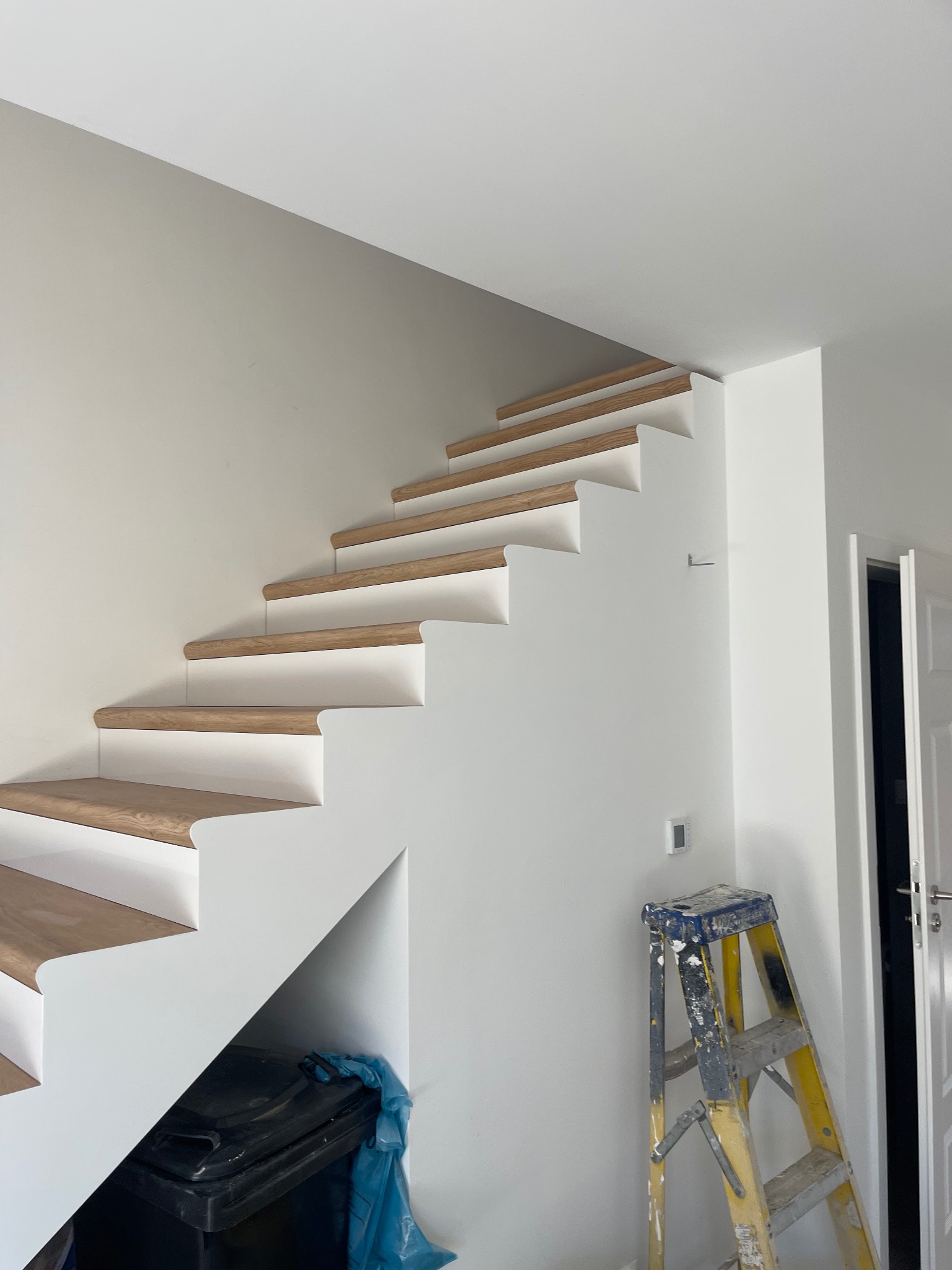 Nowoczesne, biało-drewniane schody dywanowe na białej ścianie, widoczna drabina malarska i pojemnik z folią malarską.