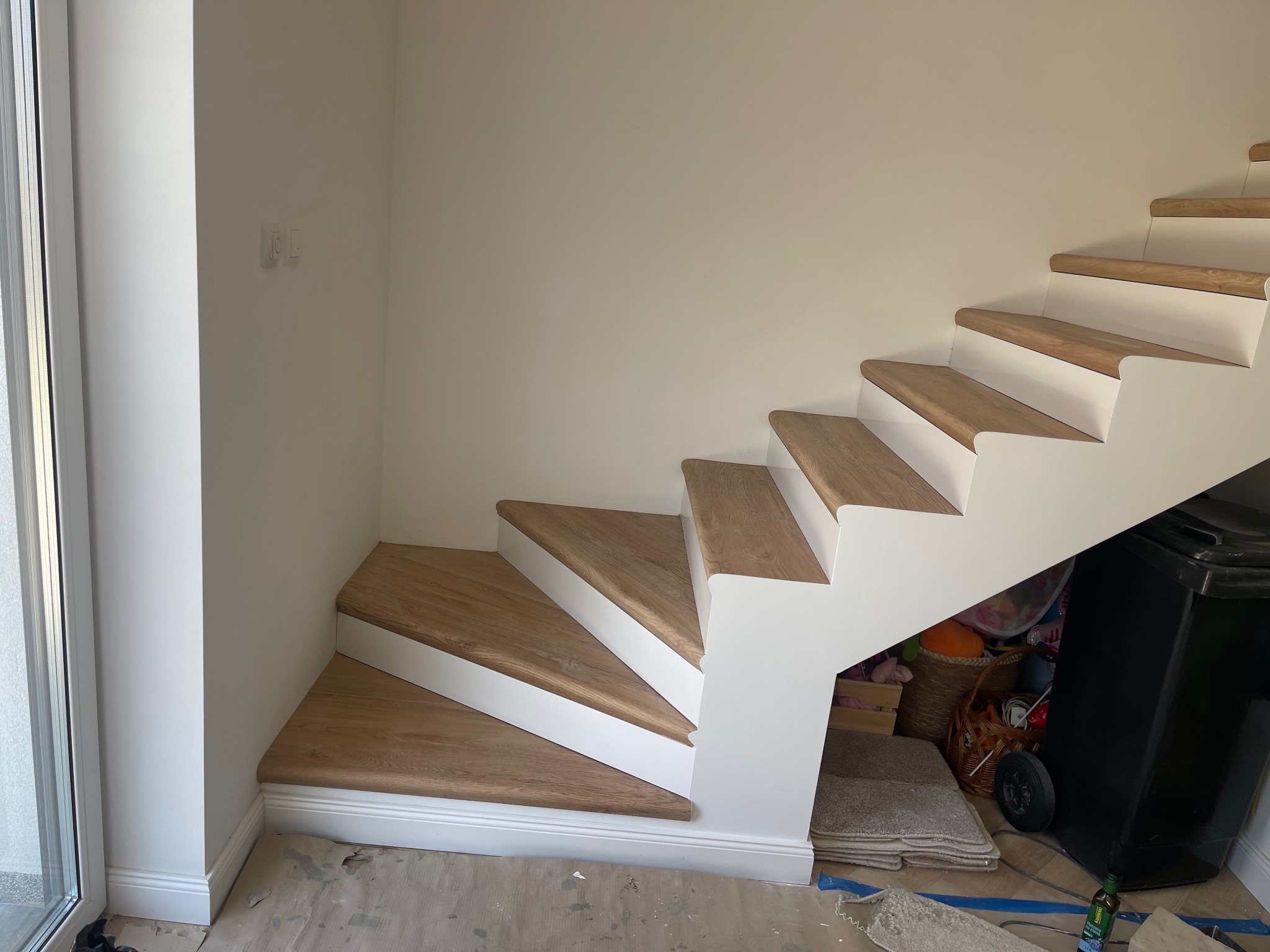 Nowe, drewniane schody z białymi podstopnicami, widok z dołu na kilka stopni, pod schodami widoczne przedmioty w nieładzie, w tle biała ściana i drzwi balkonowe.