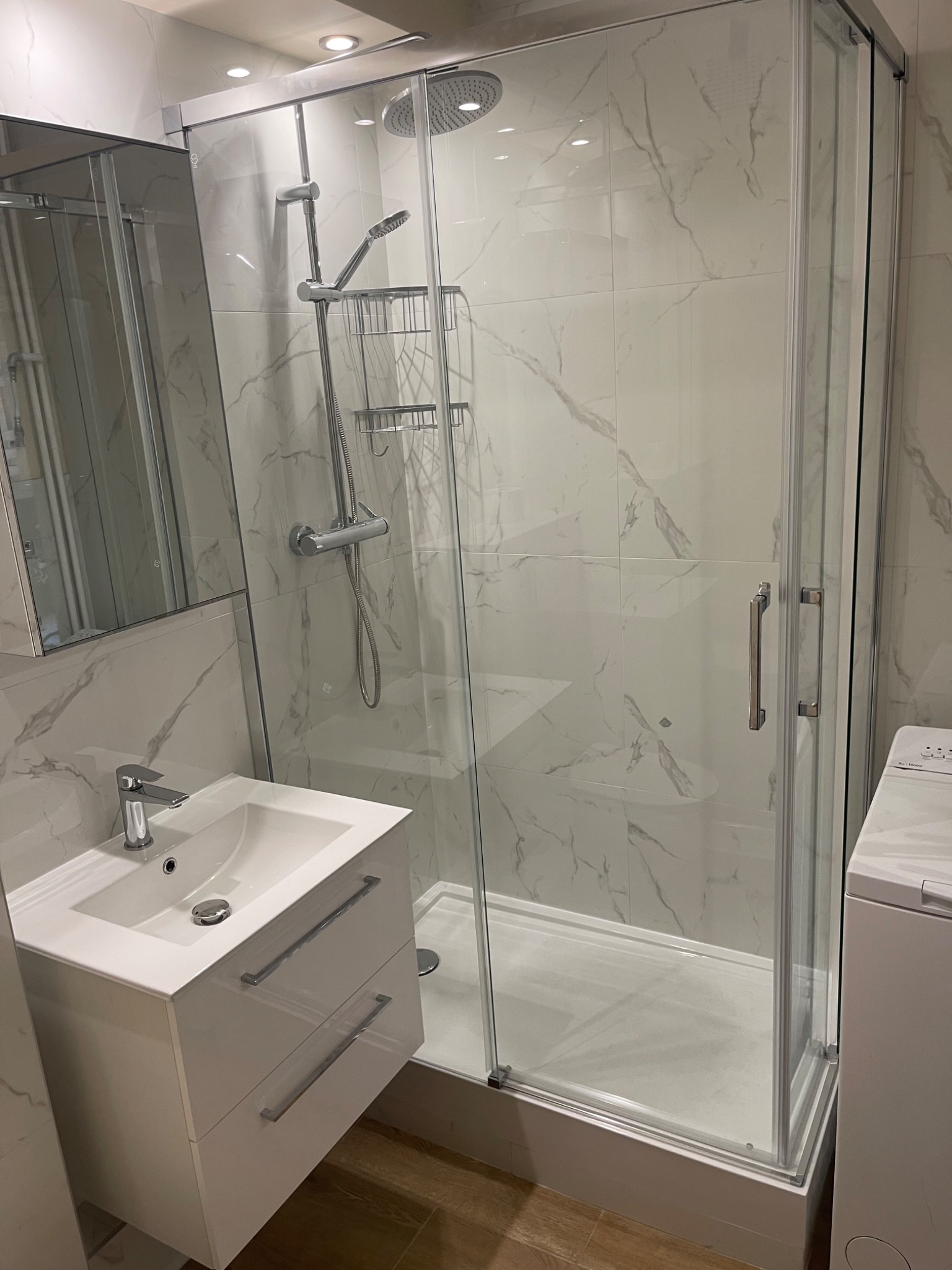 Nowoczesna łazienka z kabiną prysznicową z przezroczystego szkła, umywalką z szafką w białym połysku i marmurowymi płytkami na ścianach.