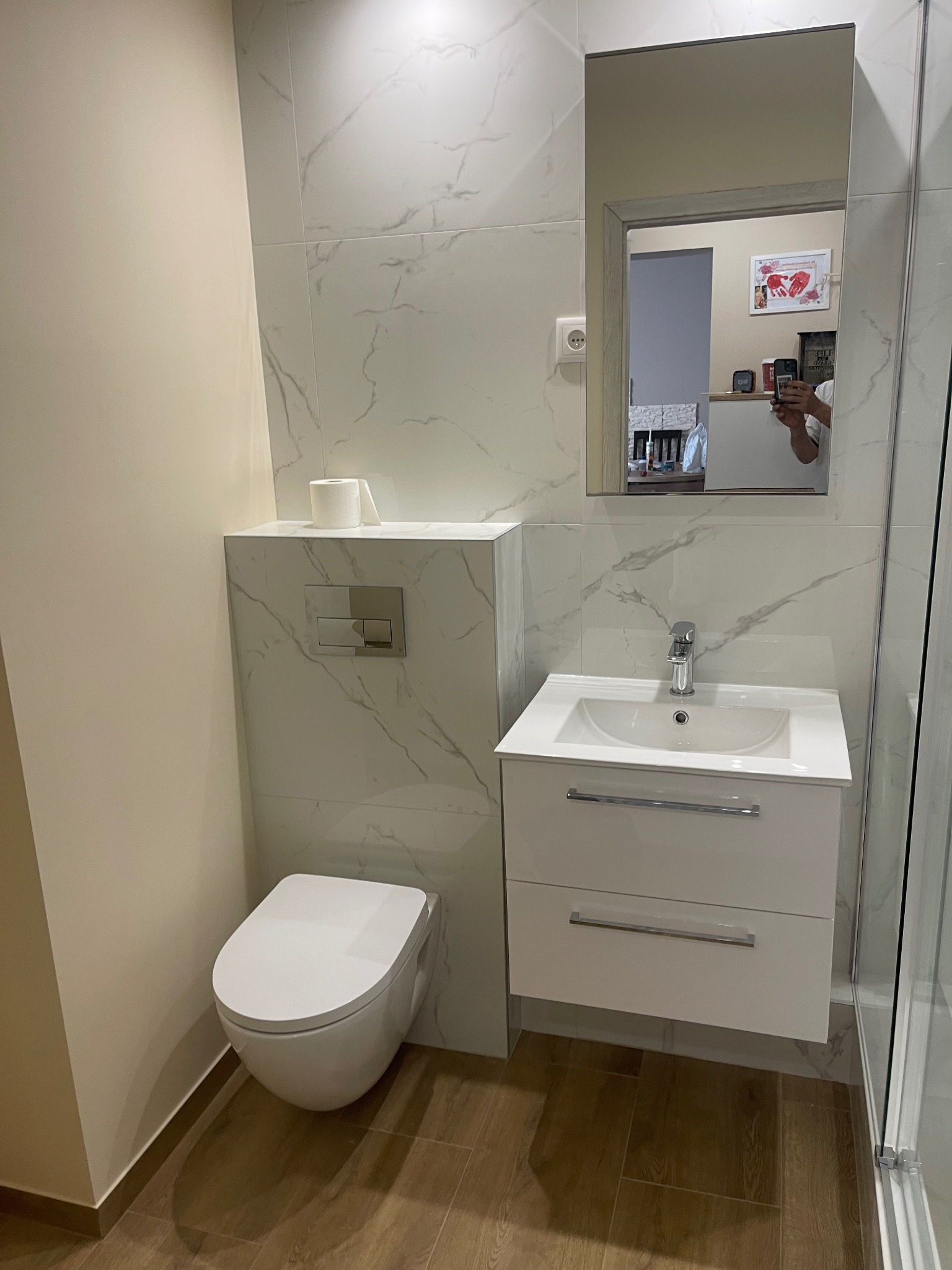 Nowoczesna łazienka z podwieszaną toaletą, białą umywalką z szafką i lustrem na ścianie wyłożonej płytkami imitującymi marmur, widoczny fragment kabiny prysznicowej.