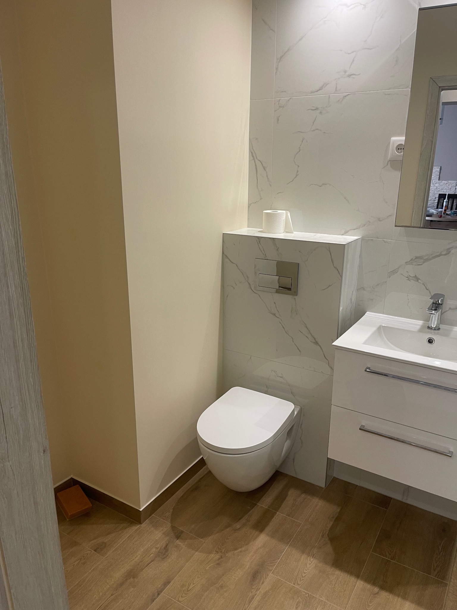 Nowoczesna łazienka po renowacji: biała ceramika, marmurowe płytki, drewnopodobne panele podłogowe, minimalistyczna zabudowa toalety z chromowanym przyciskiem spłukującym. W rogu widoczny brązowy...