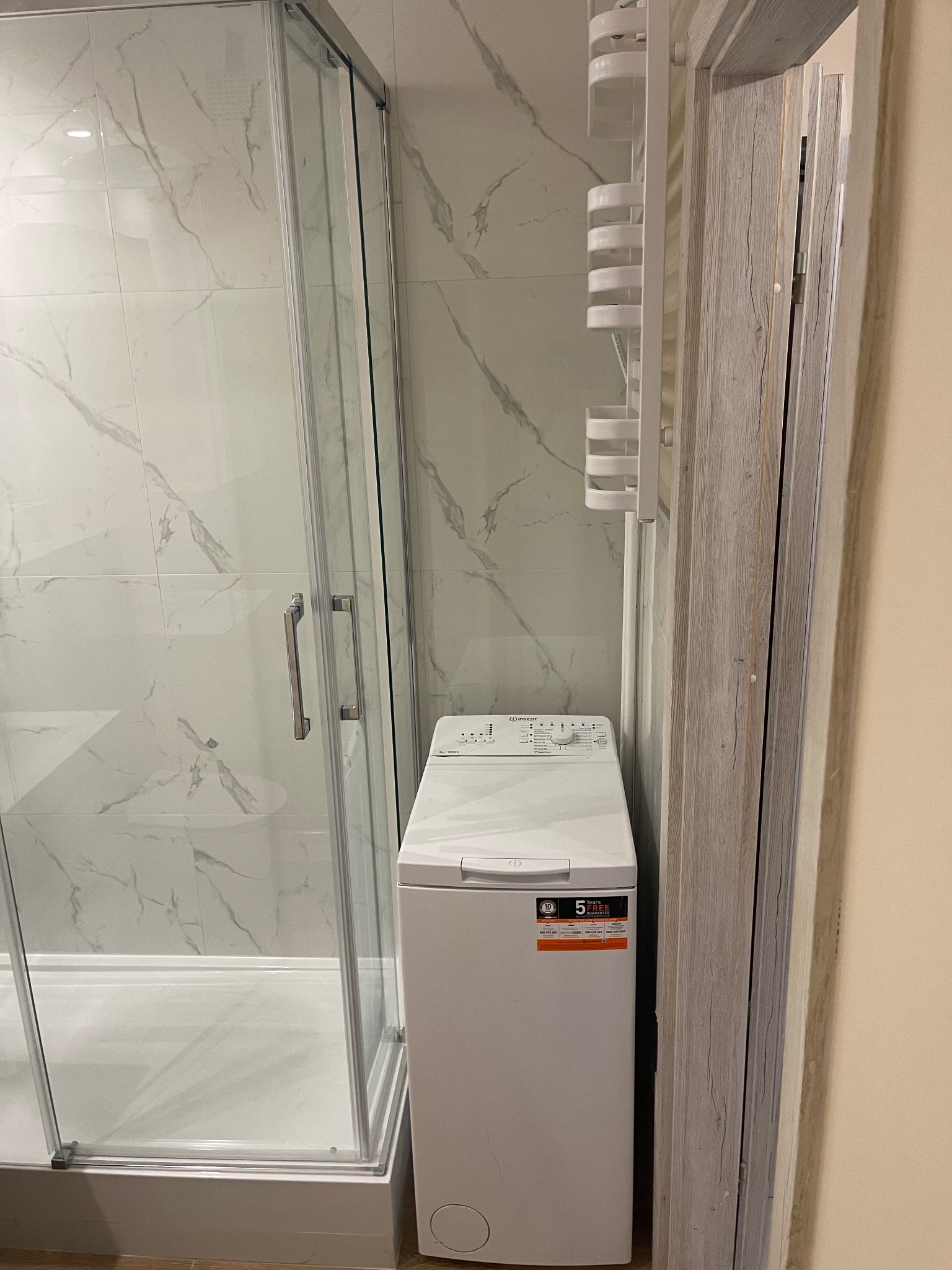 Kabina prysznicowa z białymi marmurkowymi płytkami, obok pralka i grzejnik łazienkowy, widok na wykończoną łazienkę.