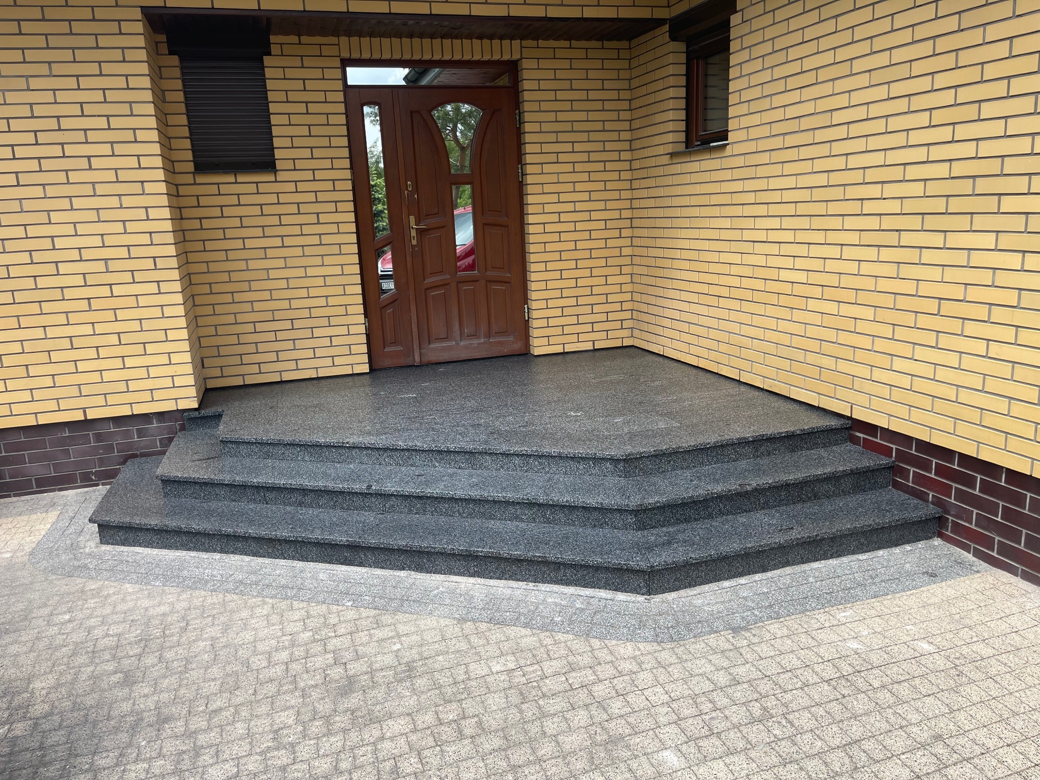 Granitowe schody wejściowe do domu z żółtej cegły, widok z dołu. Szara kostka brukowa przed schodami. Ciemnobrązowe drzwi wejściowe.