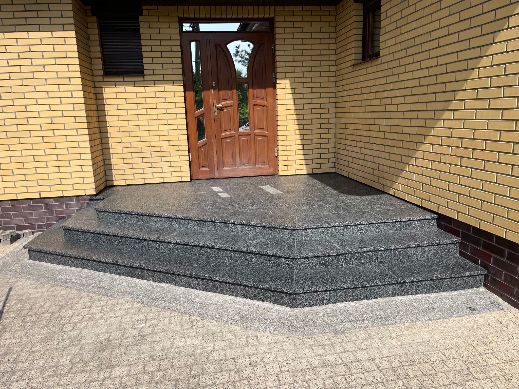 Granitowe schody wejściowe do domu z żółtej cegły, prowadzące do drewnianych drzwi z przeszkleniami. Solidne wykonanie, estetyczne wykończenie.
