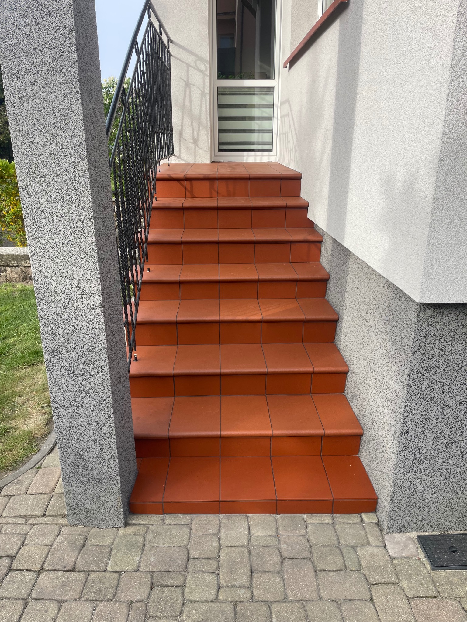 Zewnętrzne schody wyłożone terakotą, z czarną metalową balustradą i szarym filarem. Widoczna kostka brukowa na dole i fragment elewacji budynku w tle.