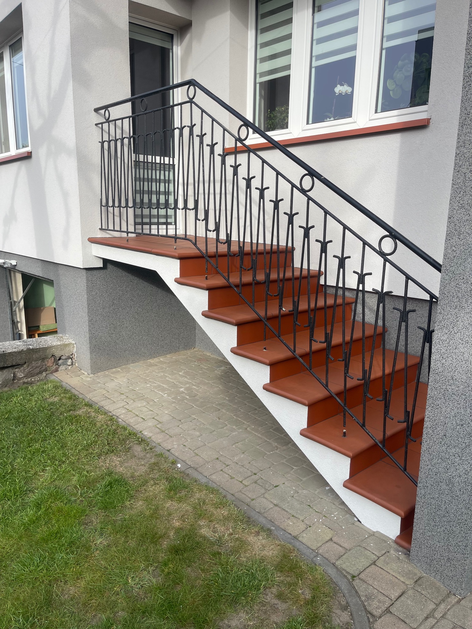 Zewnętrzne schody z terakotowymi stopniami i czarną, metalową balustradą. Widoczne okna budynku w tle i fragment trawnika. Detal wykończenia schodów.