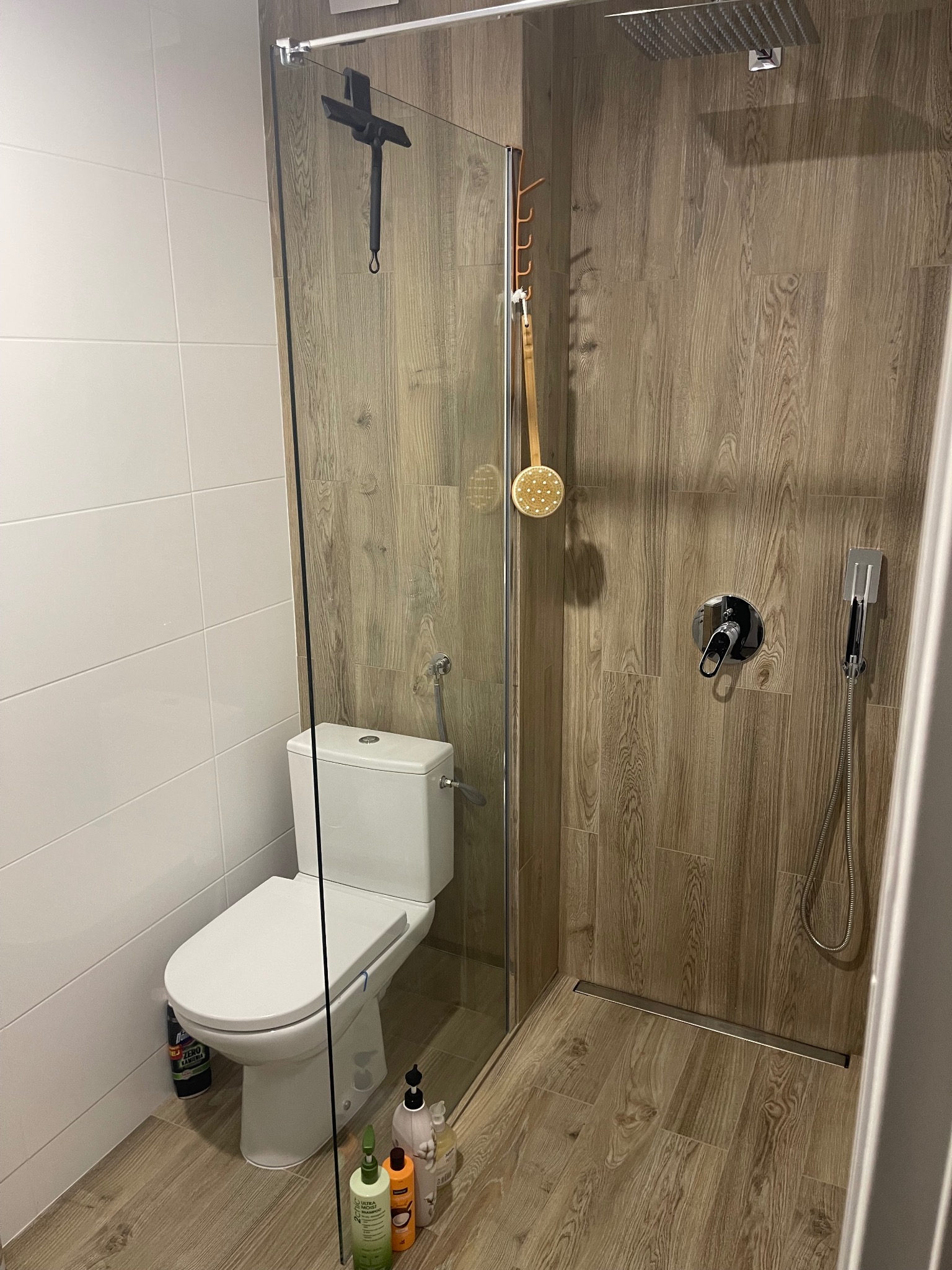 Nowoczesna łazienka po remoncie: kabina walk-in z drewnopodobnymi płytkami, toaleta i szklana ścianka. Minimalistyczny design, widoczne akcesoria łazienkowe.