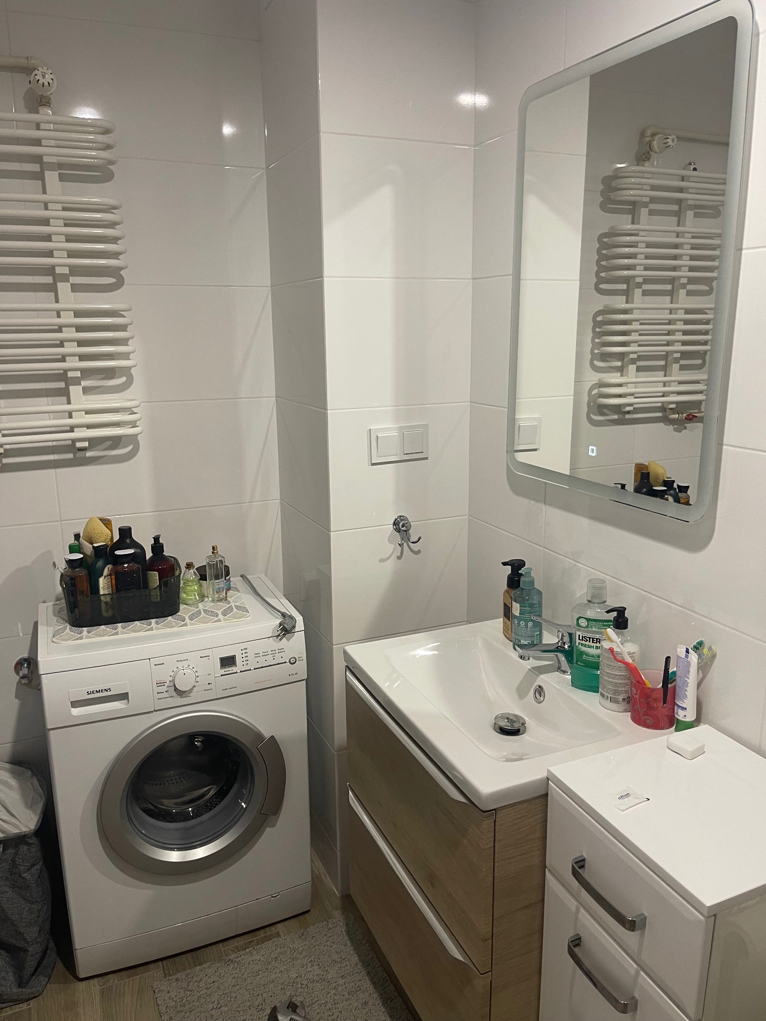 Biała łazienka po remoncie: pralka Siemens pod blatem, umywalka z szafką z drewnopodobnym frontem, lustro i grzejnik drabinkowy na tle białych płytek.