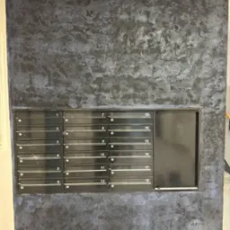 Ściana w klatce schodowej z dekoracyjną, ciemną fakturą przypominającą marmur, z zamontowanymi czarnymi skrzynkami pocztowymi z numerami od 1 do 18.