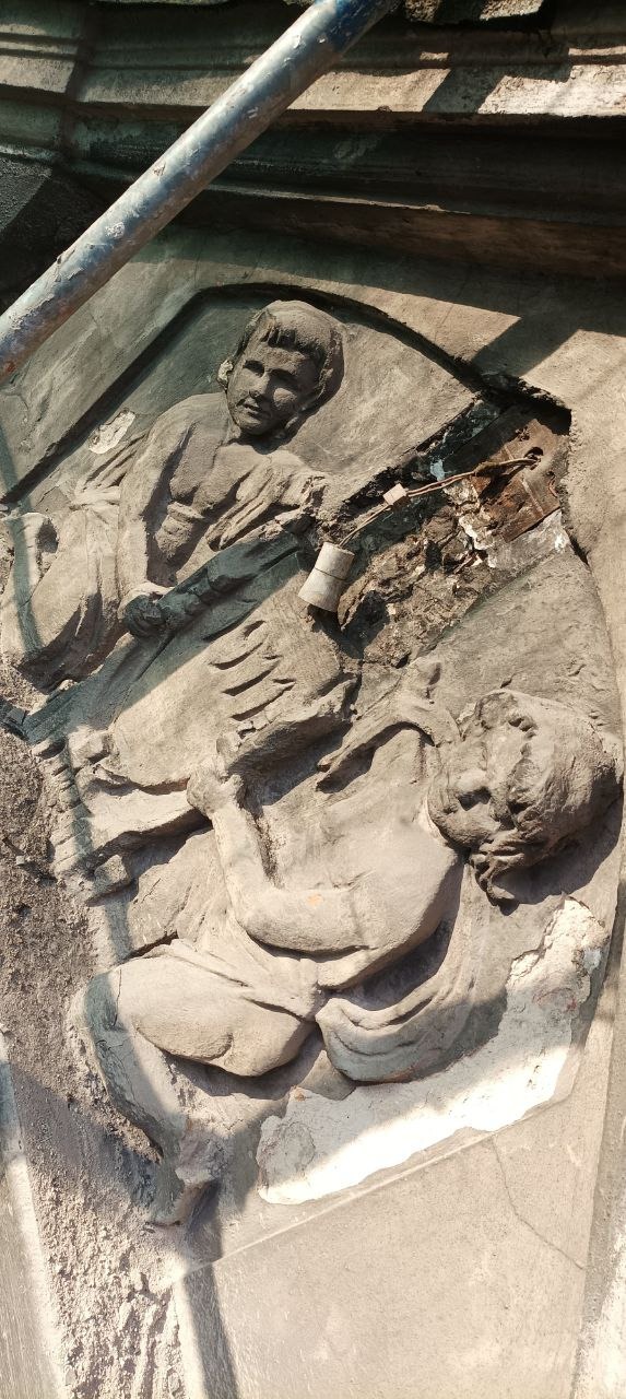 Uszkodzona płaskorzeźba z postaciami na elewacji kamienicy, widoczne ubytki i instalacja elektryczna, fragment rusztowania w górnej części kadru.