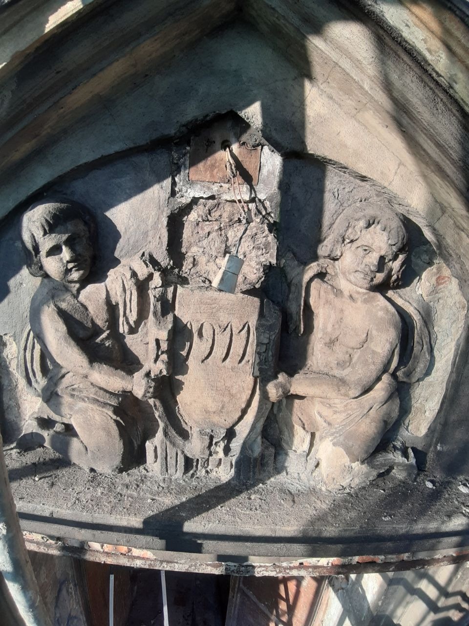 Zniszczona kamienna płaskorzeźba z dwoma aniołkami trzymającymi tarczę z numerem 1911, widoczna instalacja elektryczna, ujęcie z dołu.