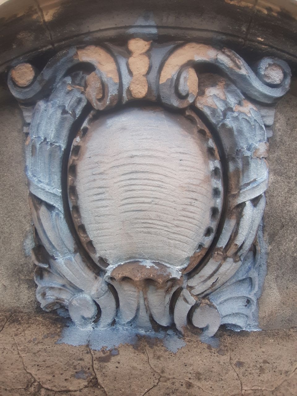 Ozdobny, owalny element architektoniczny na elewacji, z widocznymi śladami starej farby i świeżych poprawek malarskich, ukazujący detale renowacji.