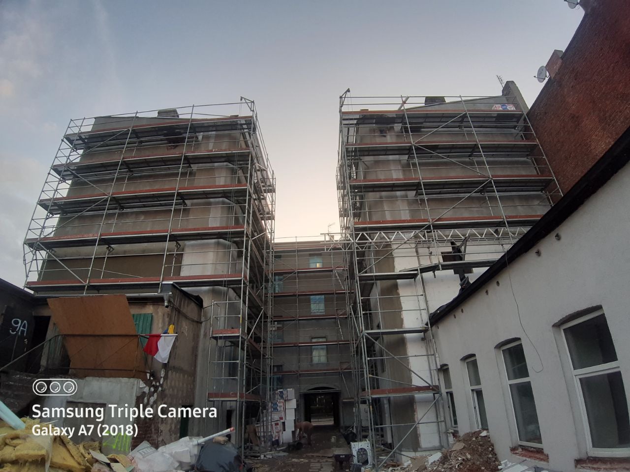 Dwa budynki w trakcie prac remontowych, otoczone rusztowaniami, widok z dołu, na dziedzińcu gruz i materiały budowlane.