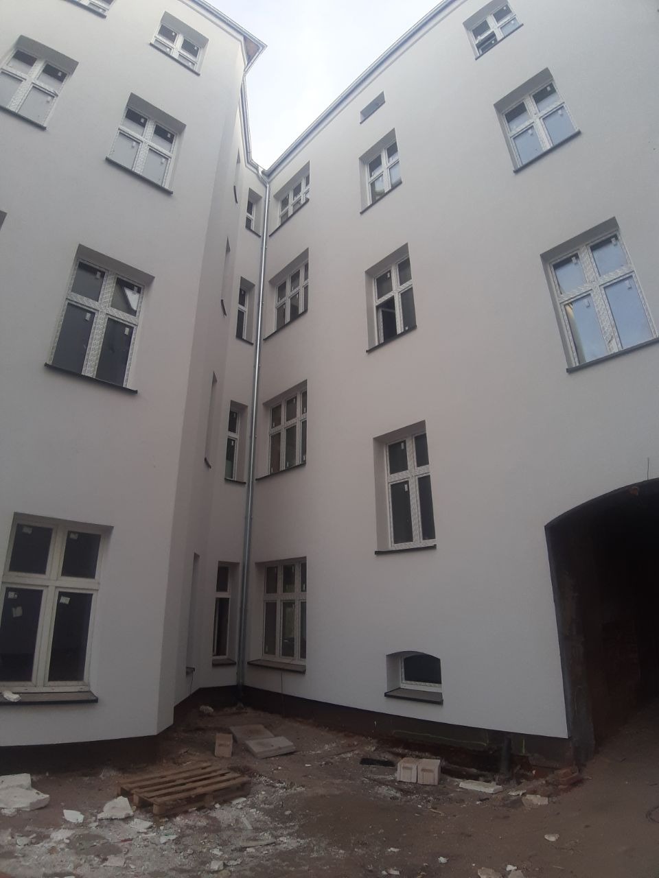 Wewnętrzny dziedziniec kamienicy po odnowieniu elewacji, widoczne nowe okna i świeżo pomalowane ściany w kolorze złamanej bieli, na ziemi resztki materiałów budowlanych.