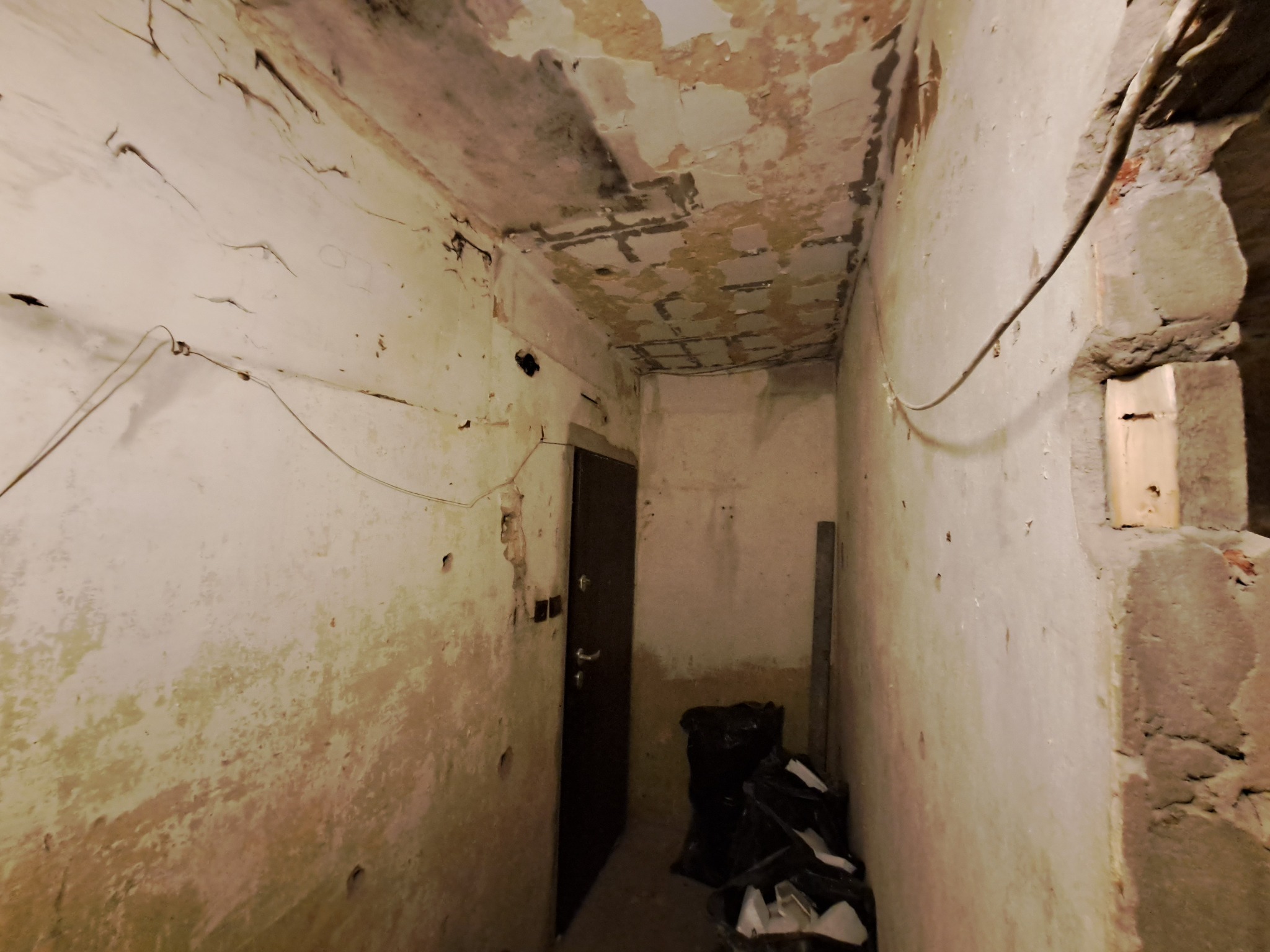 Wnętrze korytarza w trakcie prac przygotowawczych do remontu: ściany z odpadającą farbą, widoczne ślady wilgoci na suficie, worki z gruzem w rogu, surowy mur z drewnianym elementem przy wejściu.