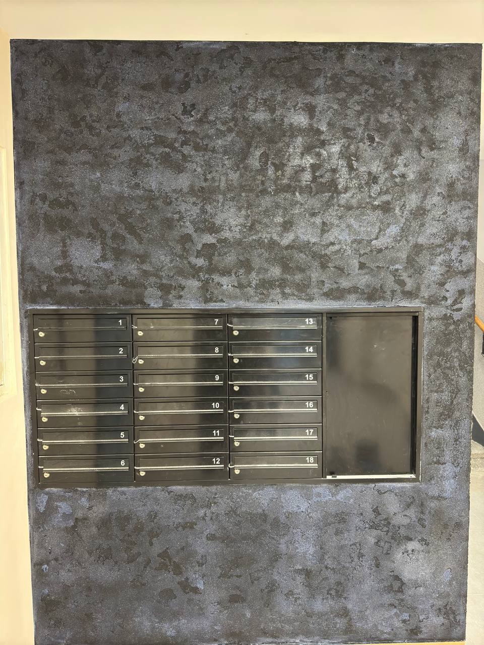 Ściana w klatce schodowej z dekoracyjną, ciemną fakturą przypominającą marmur, z zamontowanymi czarnymi skrzynkami pocztowymi z numerami od 1 do 18.