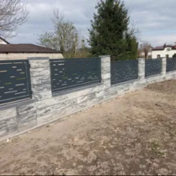 Nowoczesne ogrodzenie z paneli metalowych o geometrycznym wzorze, osadzone na podstawie z szarej kostki imitującej kamień, na tle zabudowy jednorodzinnej w pochmurny dzień.