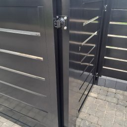 MAT - Fragment czarnej, metalowej bramy wjazdowej z poziomymi szczelinami, widoczne zawiasy i kostka brukowa na podjeździe.