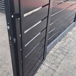 MAT - Nowoczesna, czarna brama wjazdowa z poziomymi panelami, częściowo otwarta, z widocznym zamkiem i klamką na tle kostki brukowej.