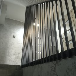 MAT - Nowoczesna balustrada schodowa z pionowych, metalowych elementów w kolorze grafitowym, zamontowana przy schodach o betonowej fakturze, widok z dołu, oświetlenie sufitowe.