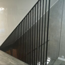 MAT - Nowoczesne schody z czarną metalową balustradą, widok z góry na szare płytki podłogowe i białą ścianę z połyskiem.