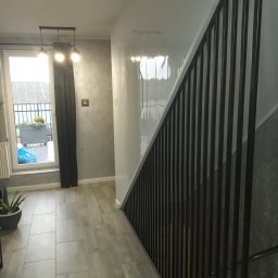 MAT - Nowoczesne schody metalowe z czarną balustradą z pionowych prętów, widok z dołu na tle szarej ściany i podłogi z płytek ceramicznych, w tle okno z widokiem na zewnątrz.