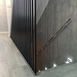 MAT - Nowoczesne schody wewnętrzne z czarną, pionową balustradą, szarą ścianą o fakturze betonu i chromowaną poręczą, oświetlone punktowo od dołu.