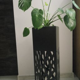 MAT - Czarna, metalowa donica o ażurowym wzorze drzewa, w której rośnie duża monstera z charakterystycznymi powycinanymi liśćmi, ustawiona w minimalistycznym wnętrzu.