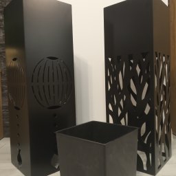 MAT - Dwa czarne, metalowe słupki dekoracyjne z wycinanymi wzorami (globus i gałęzie) oraz czarny, kwadratowy pojemnik na podłodze z paneli.