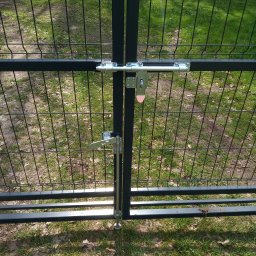 MAT - Czarna brama z metalowej siatki z dwoma różnymi typami zamków, widok z bliska na tle zieleni.