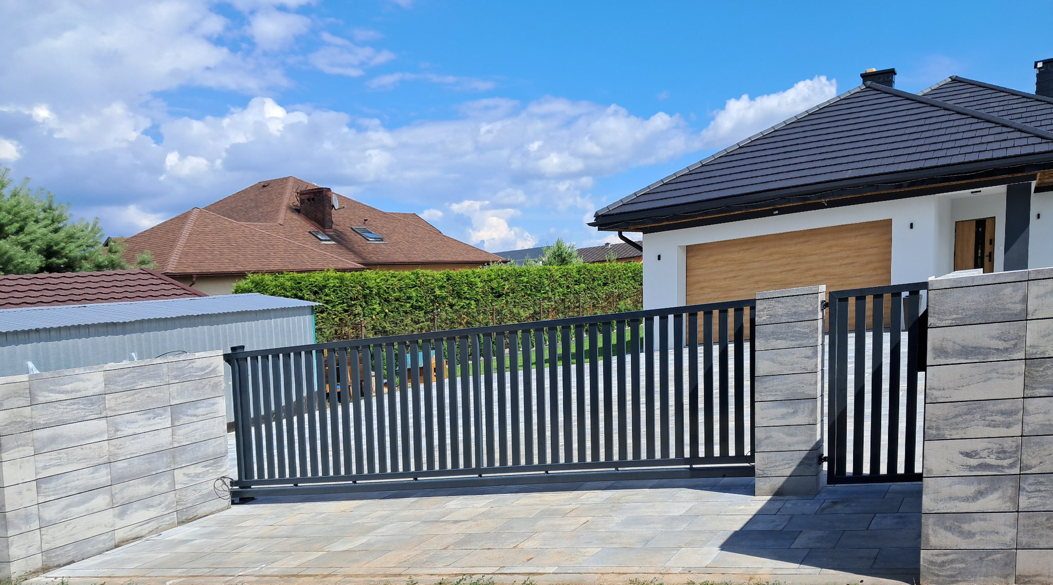 Nowoczesna przesuwna brama wjazdowa z pionowymi sztachetami w kolorze antracytowym, zamontowana na posesji z kostką brukową i ogrodzeniem z szarego kamienia łupanego, w tle dom z ciemną dachówką...