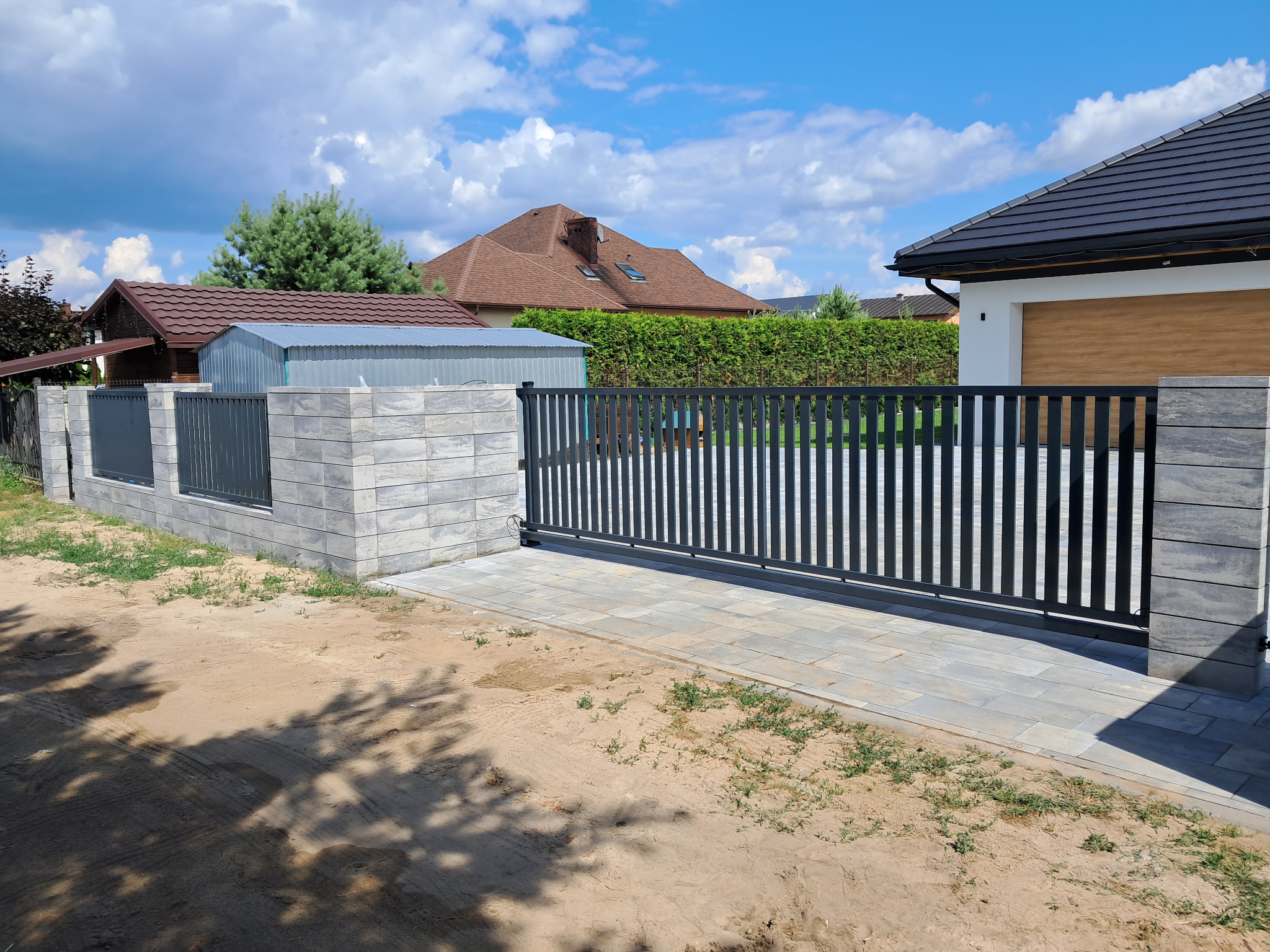 Nowoczesna, przesuwna brama wjazdowa z pionowymi szczeblami, osadzona na betonowych słupkach z szarej kostki, prowadząca na posesję z widocznym domem z brązowym dachem i garażem z drewnianą bramą.