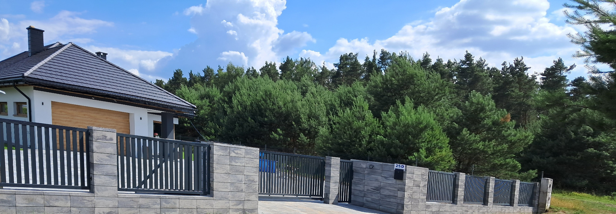 Nowoczesne ogrodzenie panelowe z podmurówką z szarej kostki, brama wjazdowa i furtka, na tle domu z ciemną dachówką i lasu iglastego.