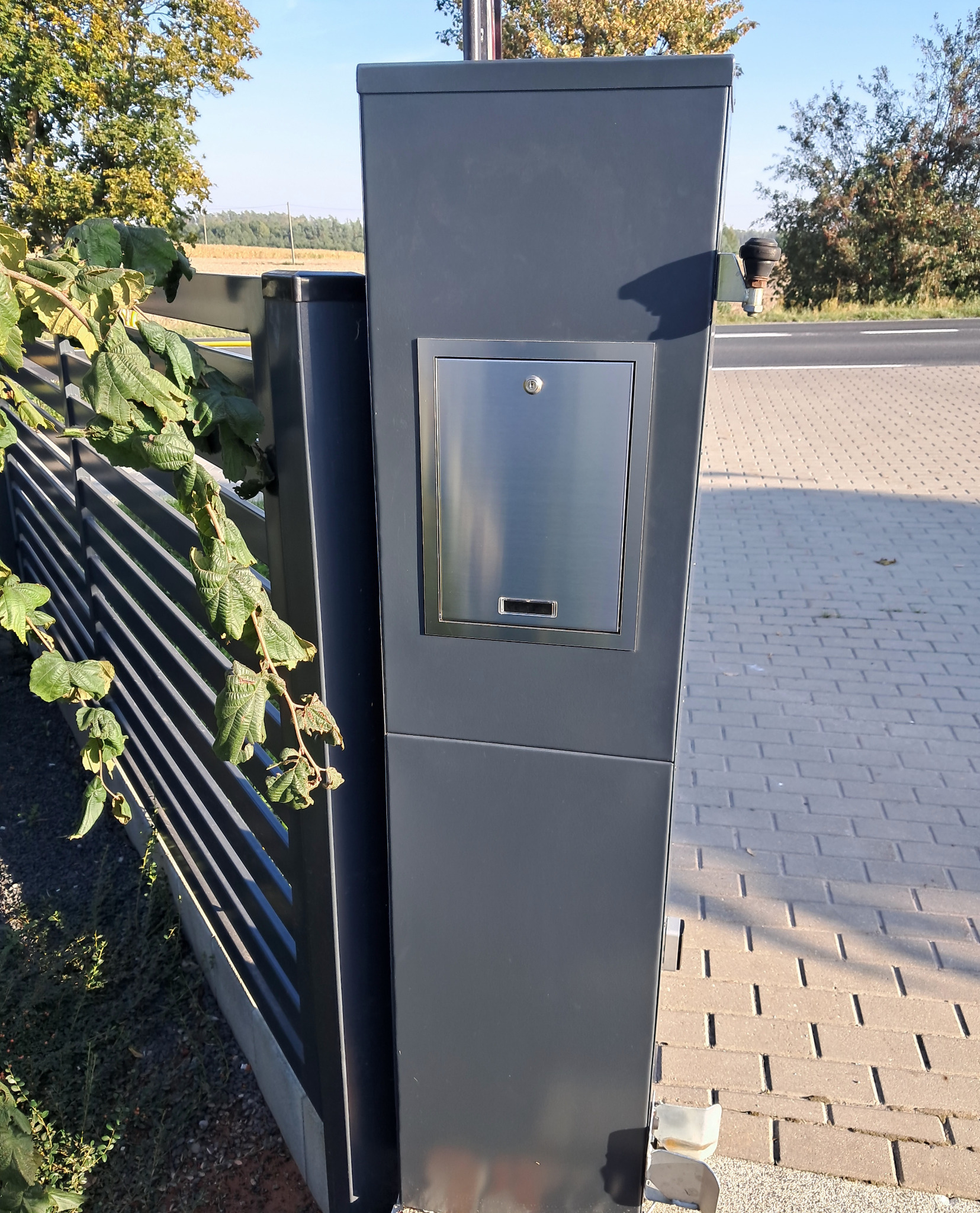 Nowoczesna brama wjazdowa z szarego metalu z wbudowaną skrzynką na listy ze stali nierdzewnej, widoczna z boku, z fragmentem ogrodzenia panelowego i zielenią w tle.