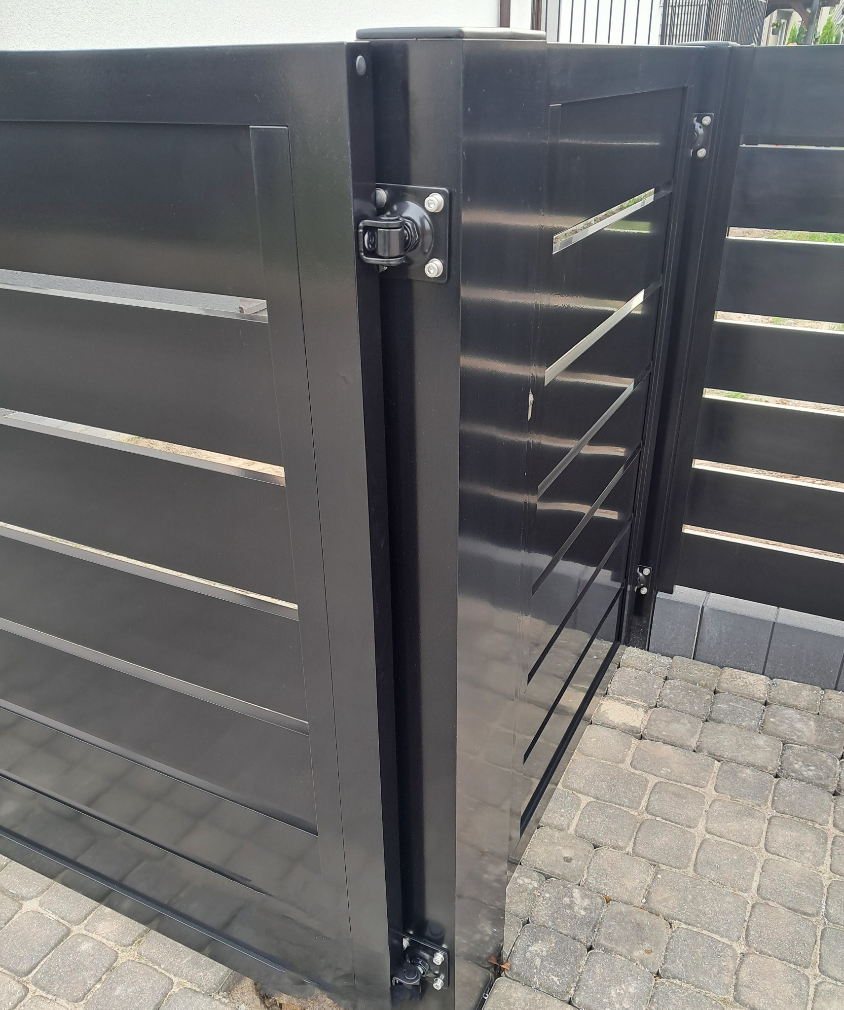 Fragment czarnej, metalowej bramy wjazdowej z poziomymi szczelinami, widoczne zawiasy i kostka brukowa na podjeździe.