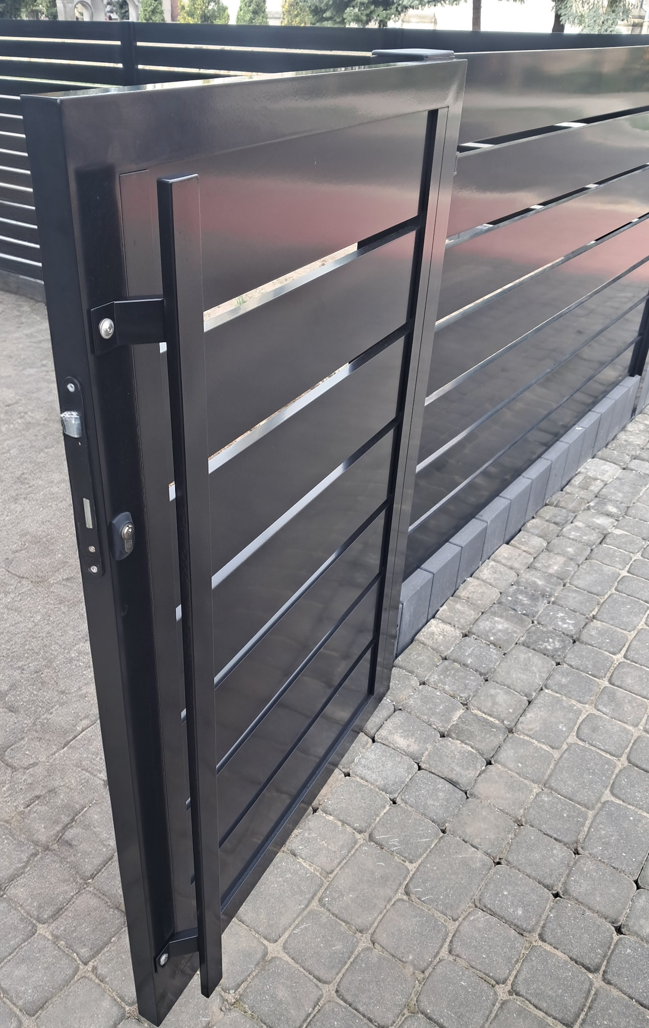 Nowoczesna, czarna brama wjazdowa z poziomymi panelami, częściowo otwarta, z widocznym zamkiem i klamką na tle kostki brukowej.