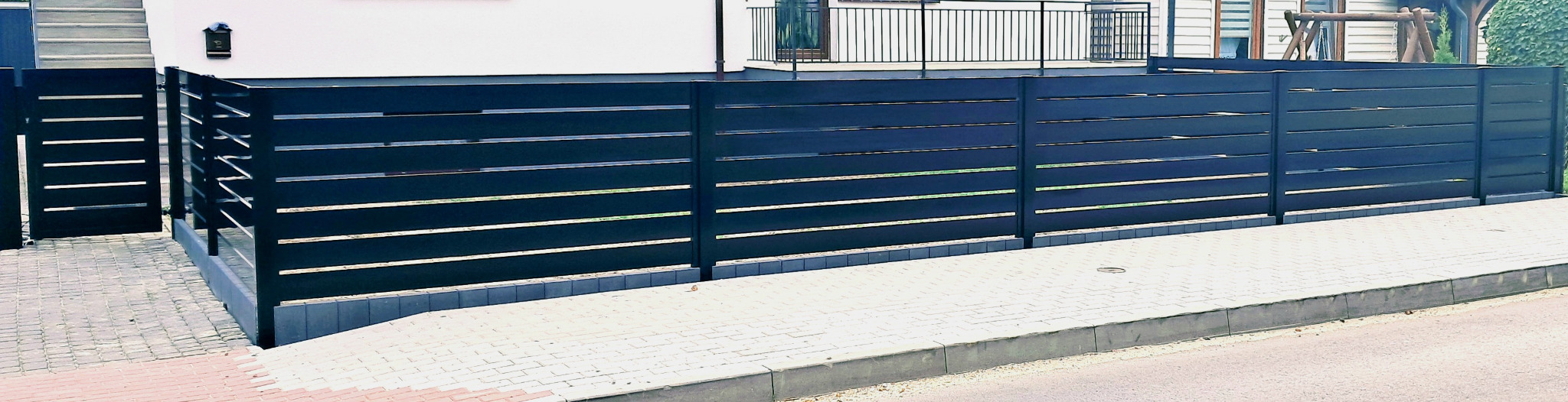 Nowoczesne, poziome ogrodzenie panelowe w kolorze antracytowym z furtką, widoczne z perspektywy ulicy, z kostką brukową w tle.