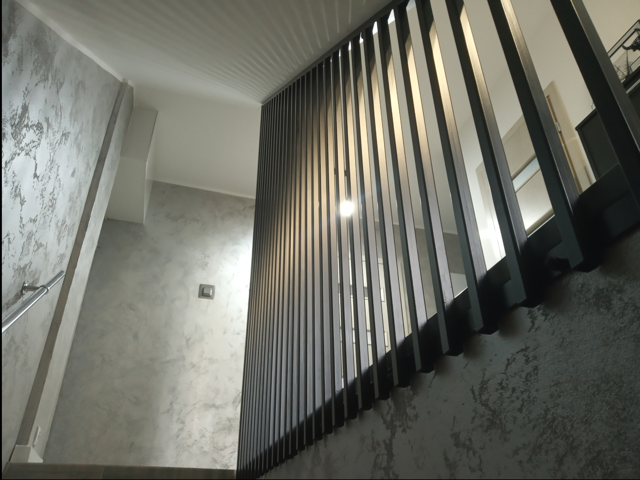 Nowoczesne schody z metalową balustradą o pionowych szczeblach, widok z dołu, ściana z dekoracyjnym tynkiem, oświetlenie sufitowe.